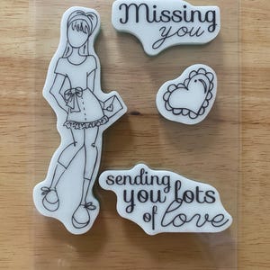 May include: A set of clear photopolymer stamps featuring a line art illustration of a person wearing a dress and holding an envelope, a scalloped heart shape, and the text phrases Missing you and sending you lots of love in black ink.