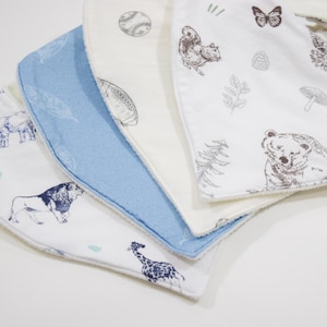Pode incluir: Uma pilha de babadores para bebês com várias ilustrações de animais e esportes. Os babadores apresentam uma paleta de cores azul, branca e creme. Os desenhos incluem um leão, uma girafa, um urso, um esquilo, beisebol e futebol americano.