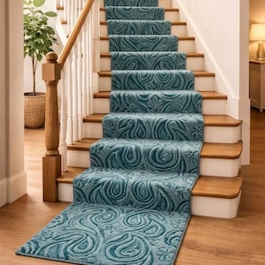 May include: Teal-colored stair runner with a repeating floral and swirl pattern. The runner covers the wooden stairs and extends onto the hardwood floor. The stairs have white risers and a wooden handrail.