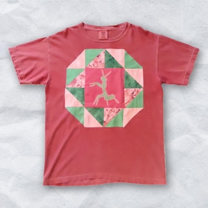 May include: A coral-colored t-shirt featuring a geometric design with a central square and surrounding triangles in shades of pink and green. A light green silhouette of a figure is in the center square. The shirt is made of soft material.