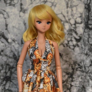 May include: A doll wearing a sleeveless dress with a cat print. The dress is orange, brown, white, and gray. The doll has blonde hair and blue eyes.