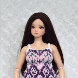 May include: A doll wearing a patterned dress with a white strap. The dress features a purple, pink, and blue geometric design. The doll has long dark hair and is wearing black flats. The background is a white, textured surface.