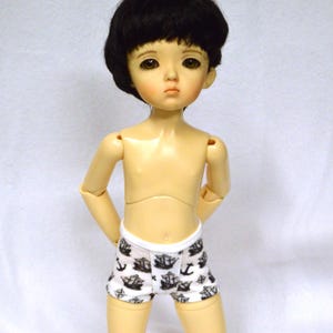 May include: A doll with a fair complexion, black hair, and wearing white and black patterned shorts. The doll has articulated joints and is standing with its arms behind its back. The shorts feature a nautical design with anchors and ships.