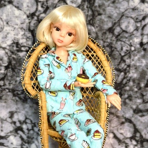 May include: A doll wearing a blue and white pajama set with a food print. The doll is sitting in a wicker chair and holding a small brown and yellow donut.