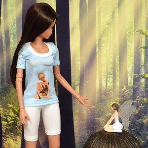Classic Smart Doll talks to fairy sitting on mushroom as she models a graphic T-shirt of an elf/fairy sitting among dandelion seeds with blue background. Jewel neckline is white ribbing. Sleeves and back are scattered tiny dandelion seeds on blue.