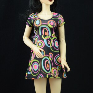 May include: A doll wearing a black and white dress with colorful circles. The dress has a scoop neck and short sleeves. The doll has long black hair and green eyes.