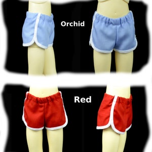 May include: Two pairs of doll shorts with white piping. The shorts on the top are orchid blue and the shorts on the bottom are red.