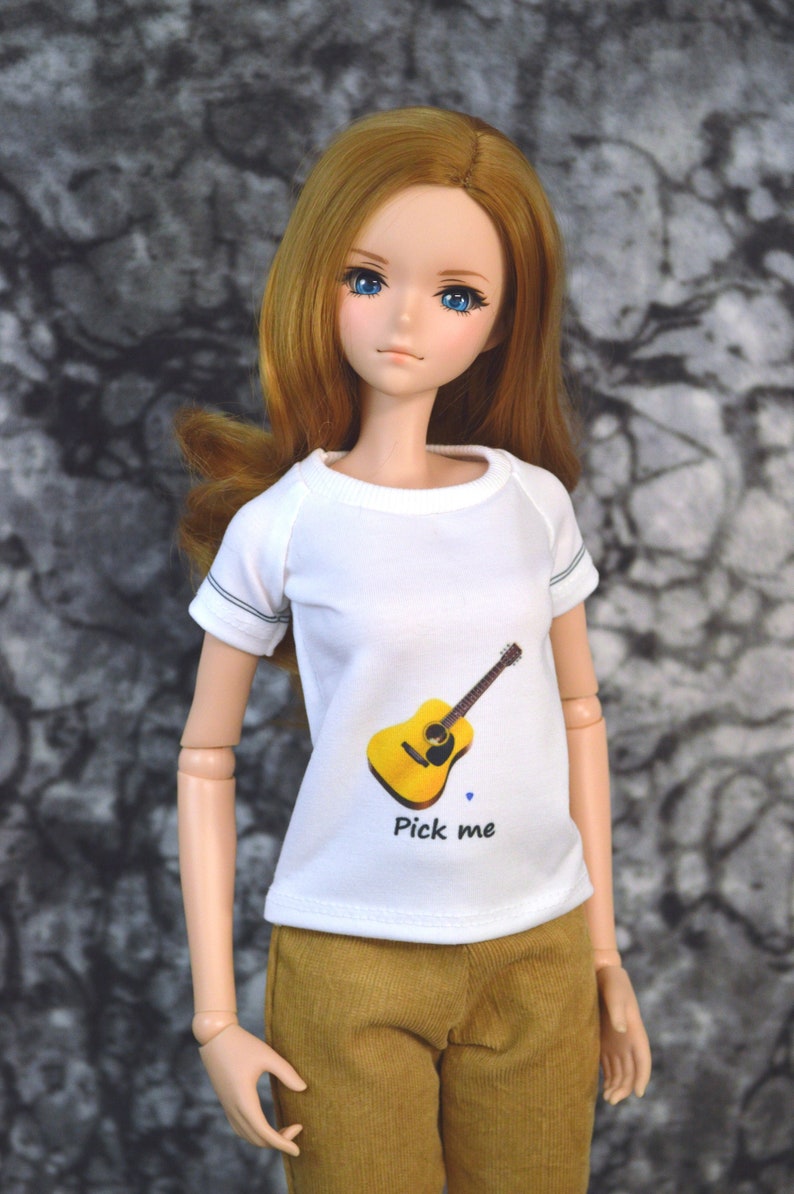 May include: A white t-shirt with a yellow guitar graphic and the text "Pick me". The doll is wearing the shirt and has long brown hair and blue eyes.