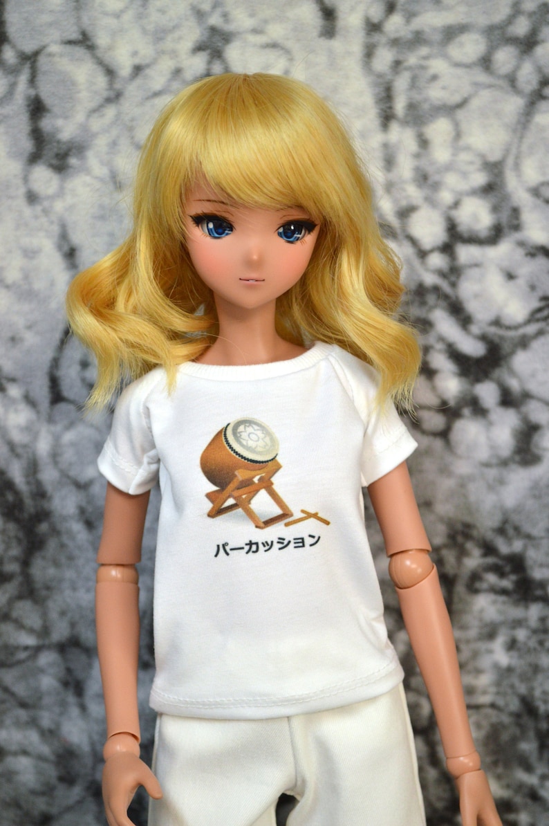 May include: A doll wearing a white t-shirt with a graphic of a Japanese drum and the text "パーカッション". The doll has long blonde hair and blue eyes.