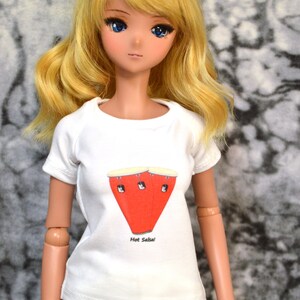May include: A doll wearing a white t-shirt with a red and white illustration of a conga drum and the text "Hot Salsa!" The doll has long blonde hair and blue eyes.
