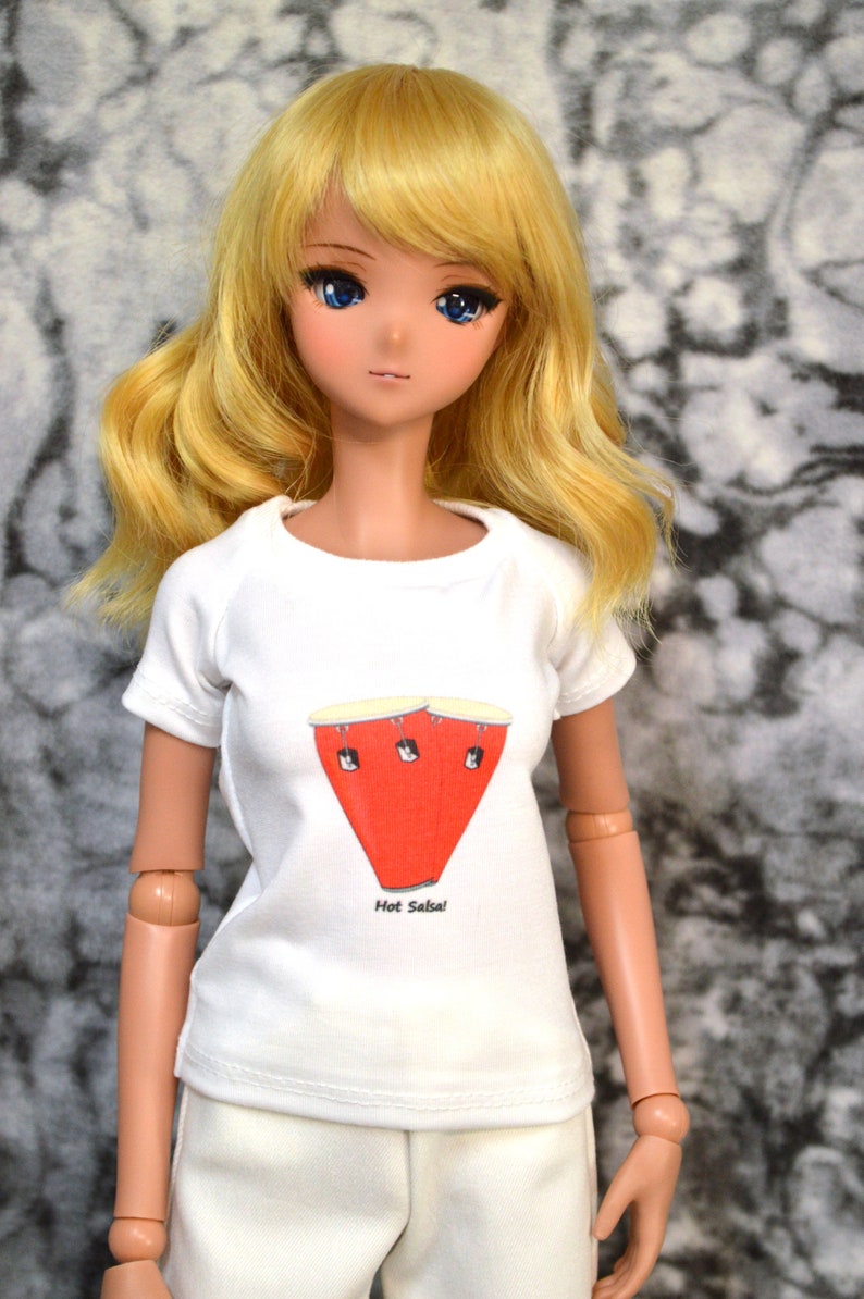 May include: A doll wearing a white t-shirt with a red and white illustration of a conga drum and the text "Hot Salsa!" The doll has long blonde hair and blue eyes.