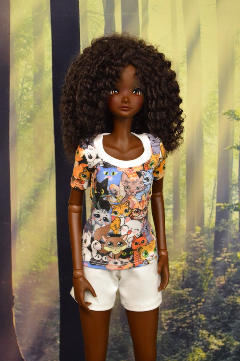 May include: A doll wearing a short-sleeved t-shirt with a white collar and white shorts. The t-shirt is covered in a colorful pattern of various cat illustrations. The doll has dark skin and curly brown hair.