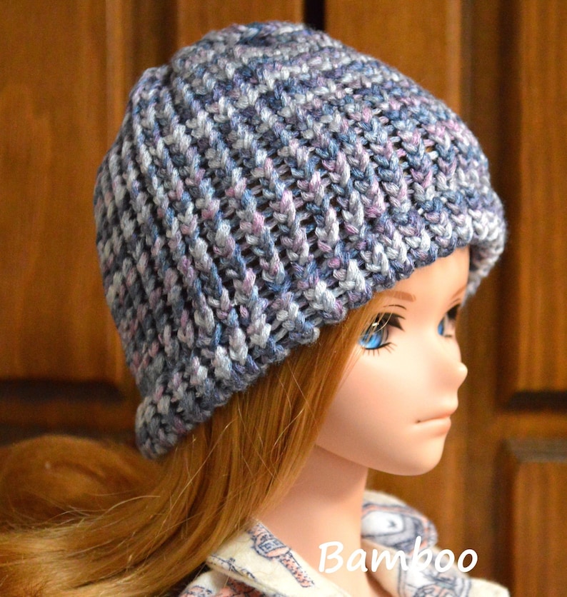 May include: A blue, grey and pink knitted beanie hat with a ribbed texture. The hat is worn by a doll with long, straight, brown hair and blue eyes.