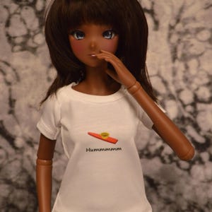 May include: A doll with brown hair and brown eyes is wearing a white t-shirt with a red and yellow kazoo graphic and the text "Hummmmmmm". The doll is also wearing white pants.
