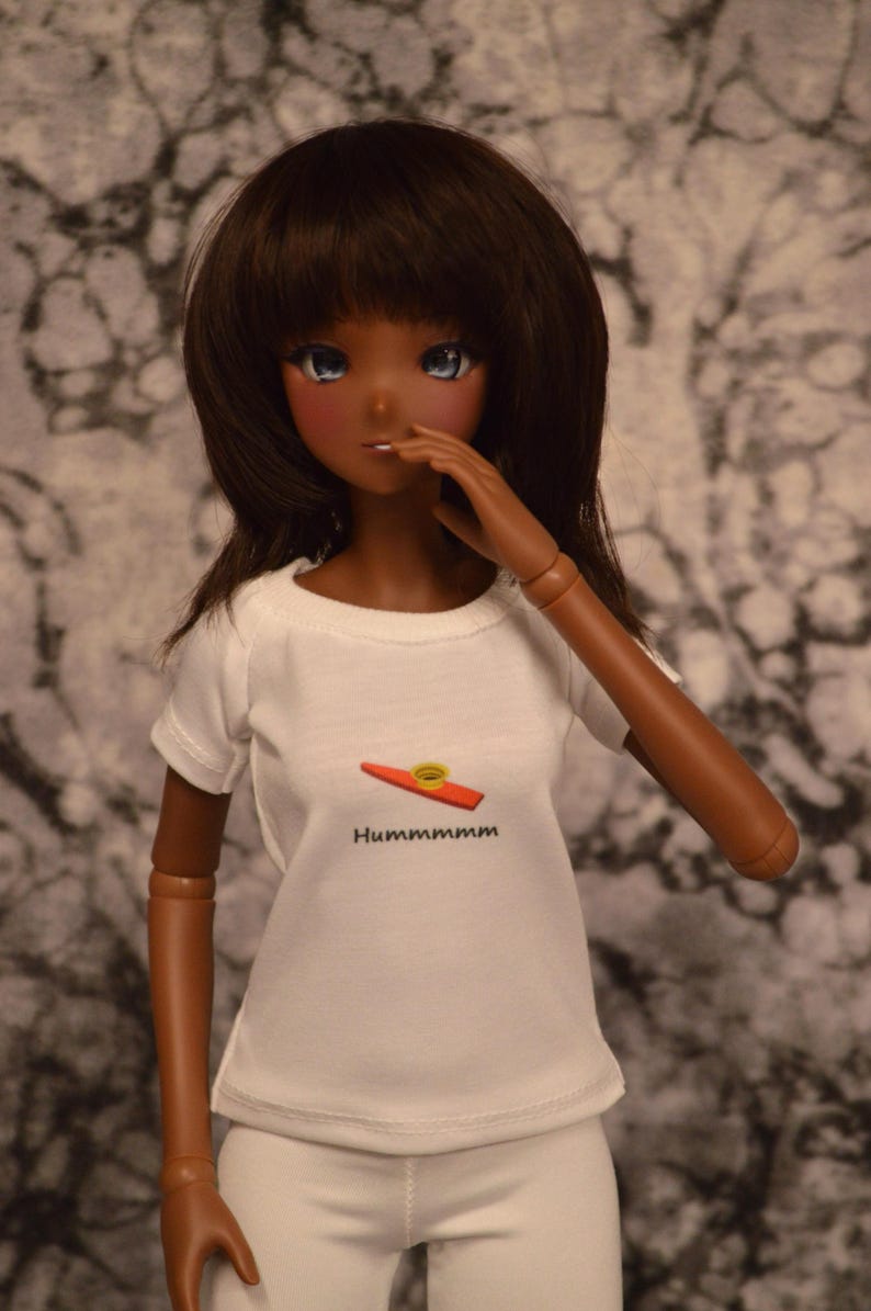 May include: A doll with brown hair and brown eyes is wearing a white t-shirt with a red and yellow kazoo graphic and the text "Hummmmmmm". The doll is also wearing white pants.