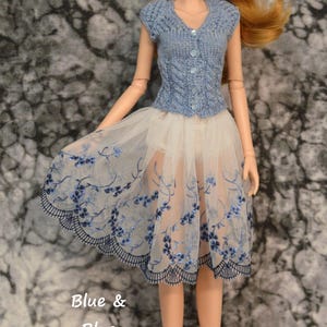 May include: A doll wearing a blue knitted vest and a white tulle skirt with blue floral embroidery. The text "Blue & Blue" is written below the doll and the text "Vest is sold separately." is written below that.