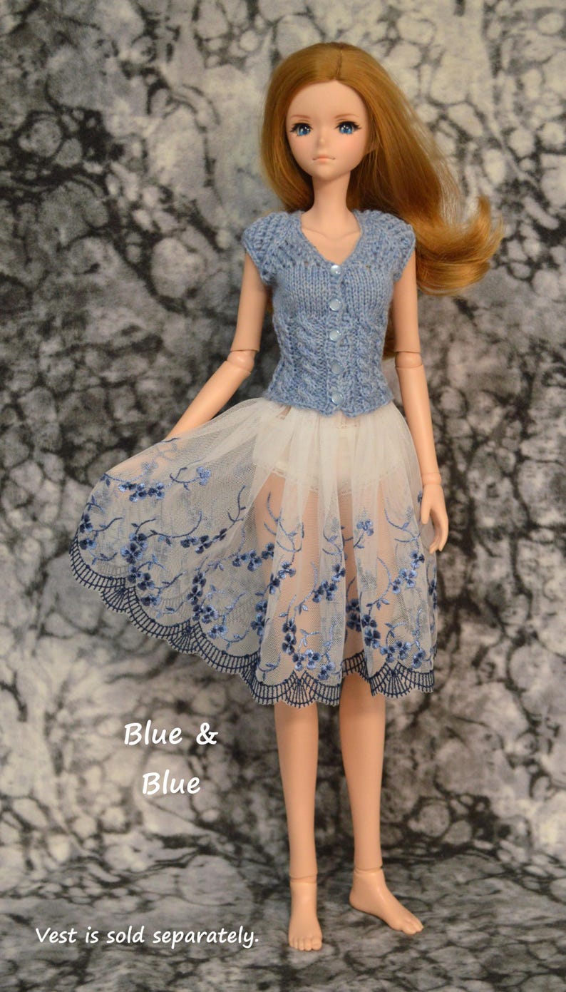May include: A doll wearing a blue knitted vest and a white tulle skirt with blue floral embroidery. The text "Blue & Blue" is written below the doll and the text "Vest is sold separately." is written below that.