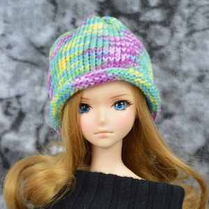 May include: A doll with long blonde hair wearing a colorful knitted hat with a brim. The hat has stripes of purple, green, yellow, and blue.