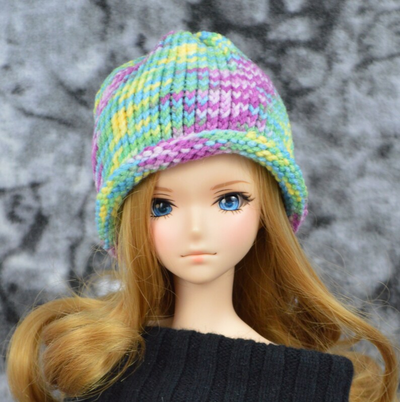 May include: A doll with long blonde hair wearing a colorful knitted hat with a brim. The hat has stripes of purple, green, yellow, and blue.