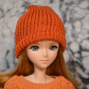 May include: A doll with long, straight, reddish-brown hair wearing a bright orange knitted beanie hat. The doll has blue eyes and a neutral expression.