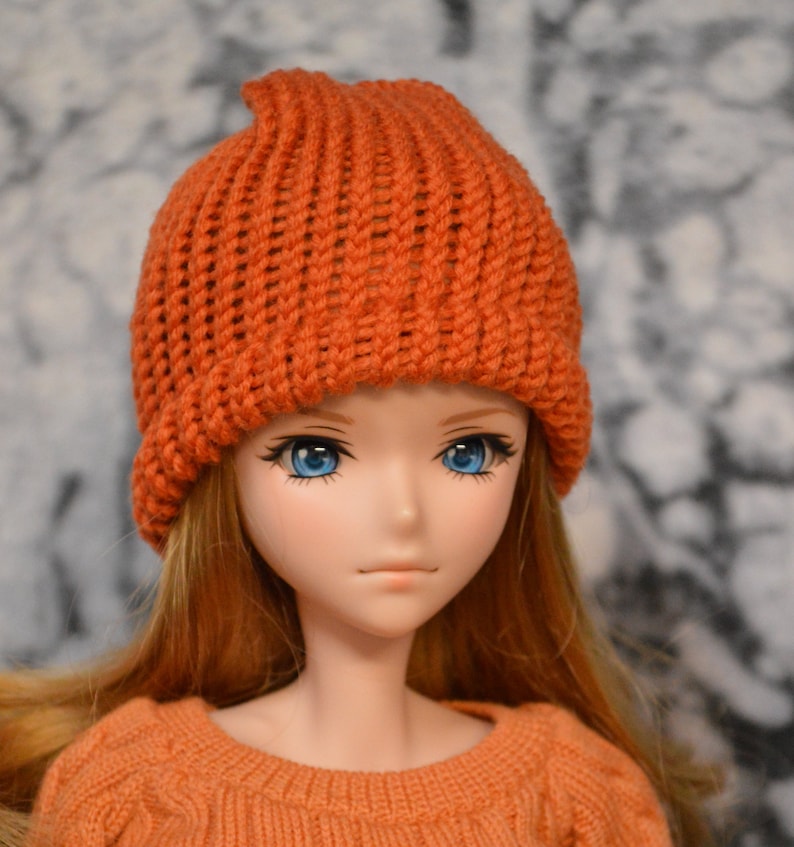 May include: A doll with long, straight, reddish-brown hair wearing a bright orange knitted beanie hat. The doll has blue eyes and a neutral expression.