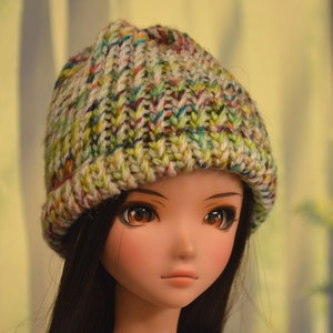 May include: A doll with long brown hair wearing a white, green and purple knitted beanie hat.