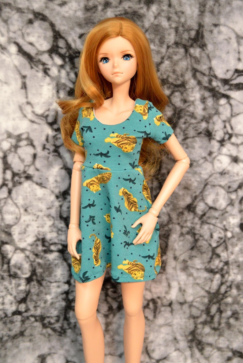 Smart Doll models a short skater dress of a stretchy knit fabric decorated with tigers on a green background. Details include a jewel neckline and short sleeves. Bodice is lined in white as it hugs her body closely.