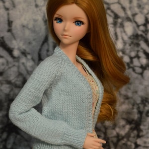 May include: A doll with long, wavy, auburn hair wearing a light blue knitted cardigan over a floral dress.