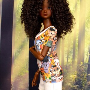 May include: A doll with dark skin and curly brown hair is wearing a white and colorful cat-print top and white shorts. The doll is posed in front of a blurred background of trees and sunlight.