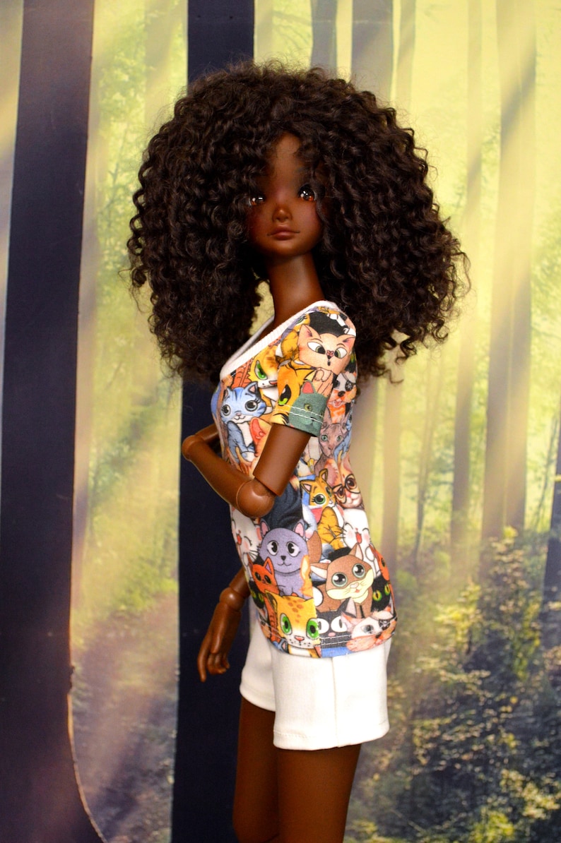 May include: A doll with dark skin and curly brown hair is wearing a white and colorful cat-print top and white shorts. The doll is posed in front of a blurred background of trees and sunlight.