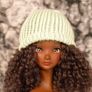 May include: A doll with brown curly hair wearing a light green knitted beanie.