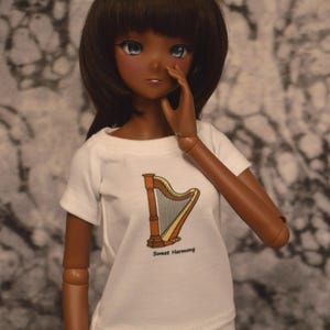 May include: A doll with brown skin and long brown hair is wearing a white t-shirt with a harp graphic and the text "Sweet Harmony". The doll is wearing white pants and has blue eyes.