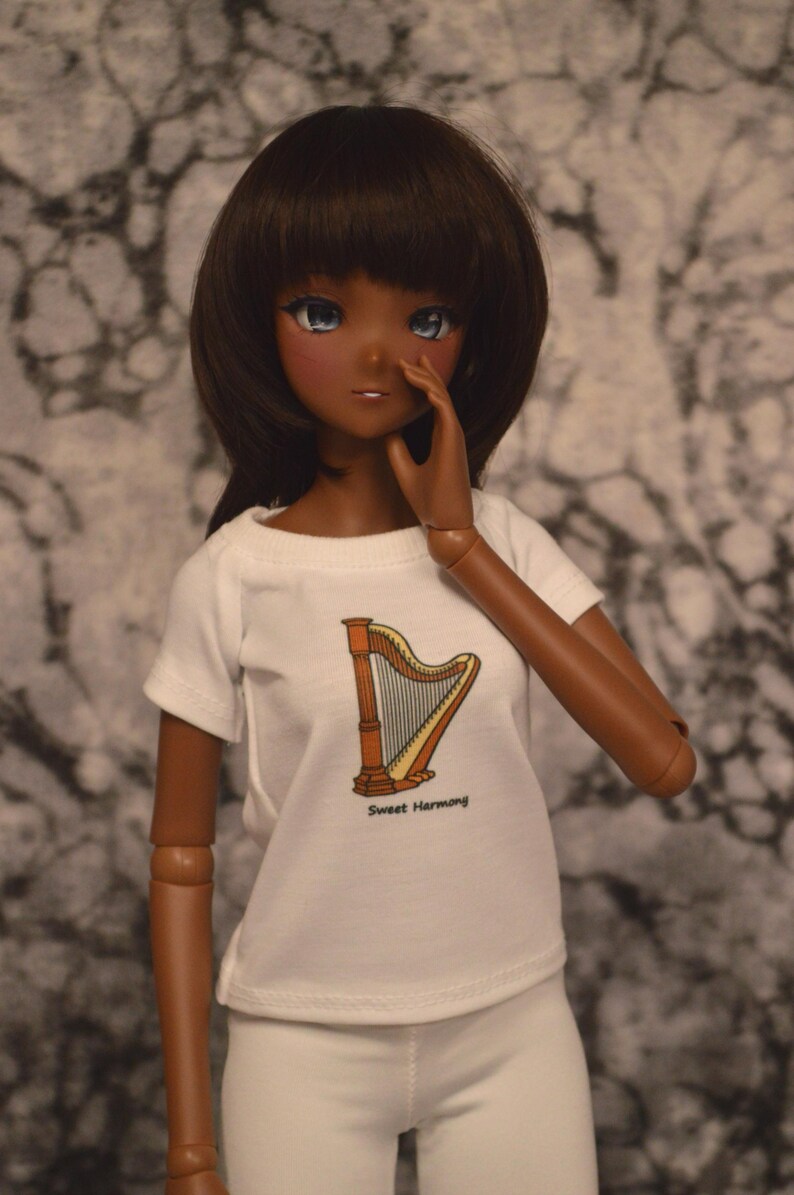 May include: A doll with brown skin and long brown hair is wearing a white t-shirt with a harp graphic and the text "Sweet Harmony". The doll is wearing white pants and has blue eyes.