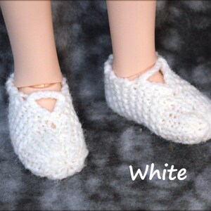 May include: White knitted doll shoes with a criss-cross strap detail. The shoes are on a gray textured surface. White.