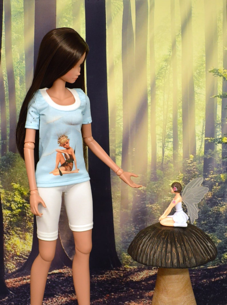 Classic Smart Doll talks to fairy sitting on mushroom as she models a graphic T-shirt of an elf/fairy sitting among dandelion seeds with blue background. Jewel neckline is white ribbing. Sleeves and back are scattered tiny dandelion seeds on blue.