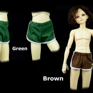 May include: Two pairs of doll-sized athletic shorts, one green and one brown, with white trim. The shorts have a retro style.