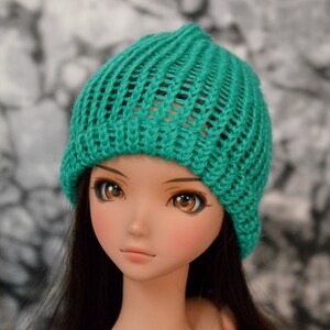 May include: A doll with long brown hair wearing a teal knitted beanie hat.
