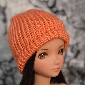 May include: A close-up of a doll wearing a knitted beanie hat in a peachy orange colour. The hat has a ribbed texture and is pulled down over the doll's forehead.