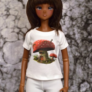 Może przedstawiać: Lalka ubrana w białą koszulkę z nadrukiem domu z grzyb&oacute;w i białe spodnie. Na koszulce widnieje napis "My Mushroom Home".