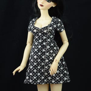 May include: A doll with black hair and green eyes wearing a black and white patterned dress.