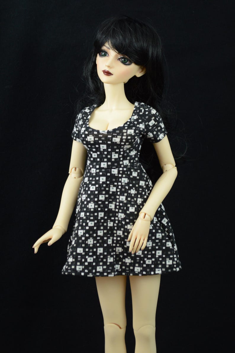 May include: A doll with black hair and green eyes wearing a black and white patterned dress.