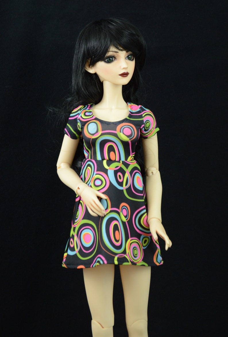 May include: A doll wearing a black and white dress with colorful circles. The dress has a scoop neck and short sleeves. The doll has long black hair and green eyes.