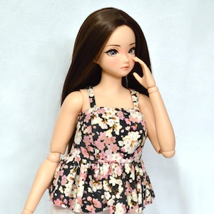 May include: A doll wearing a black and pink floral print top with a ruffled hem and thin straps. The doll has long brown hair and blue eyes.