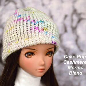 May include: A white knitted hat with a colourful yarn blend. The hat has a ribbed texture and a slouchy fit. The hat is being worn by a doll with brown hair and brown eyes. The text "Cake Pop: Cashmere/Merino Blend" is visible in the image.
