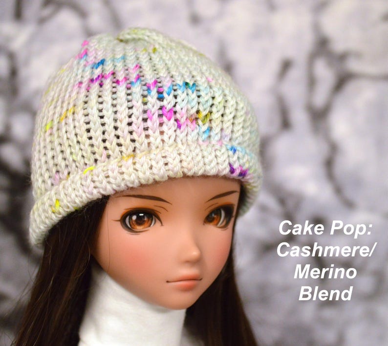 May include: A white knitted hat with a colourful yarn blend. The hat has a ribbed texture and a slouchy fit. The hat is being worn by a doll with brown hair and brown eyes. The text "Cake Pop: Cashmere/Merino Blend" is visible in the image.