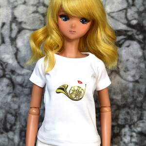 May include: A doll with long blonde hair wearing a white t-shirt with a gold french horn graphic and white pants.