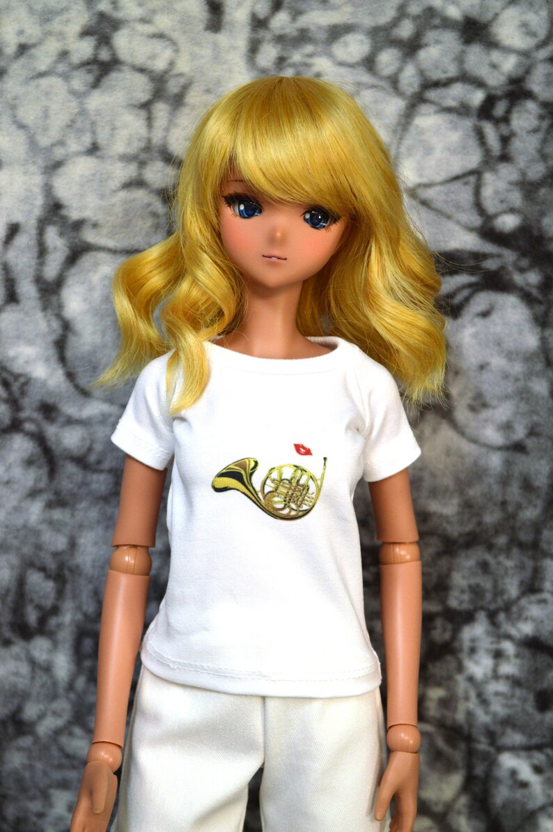 May include: A doll with long blonde hair wearing a white t-shirt with a gold french horn graphic and white pants.