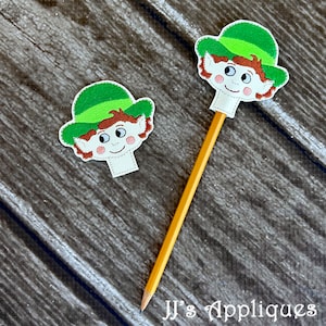 May include: Two felt elf finger puppets with green hats and brown hair. The puppet on the left is laying flat on a wooden surface. The puppet on the right is attached to a yellow pencil.