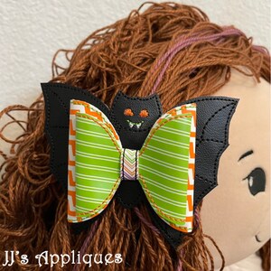 May include: A black bat-shaped hair bow with green and orange striped fabric and a stitched-on bat face.