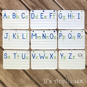 May include: Nine white cards with blue and green embroidered letters of the alphabet, A through Z, on each card. The cards are arranged in three rows of three cards each. The last card has a graduation cap and tassel embroidered on it. The cards are on a wooden surface. The text "JJ's Appliques" is at the bottom of the image.